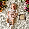 baby, infant, smiling, gingham, dress, shoes, socks, blanket, pattern, flowers, floral_arrangement, wooden_sign, months, cute, happy, lying_down, portrait, indoor, child, person