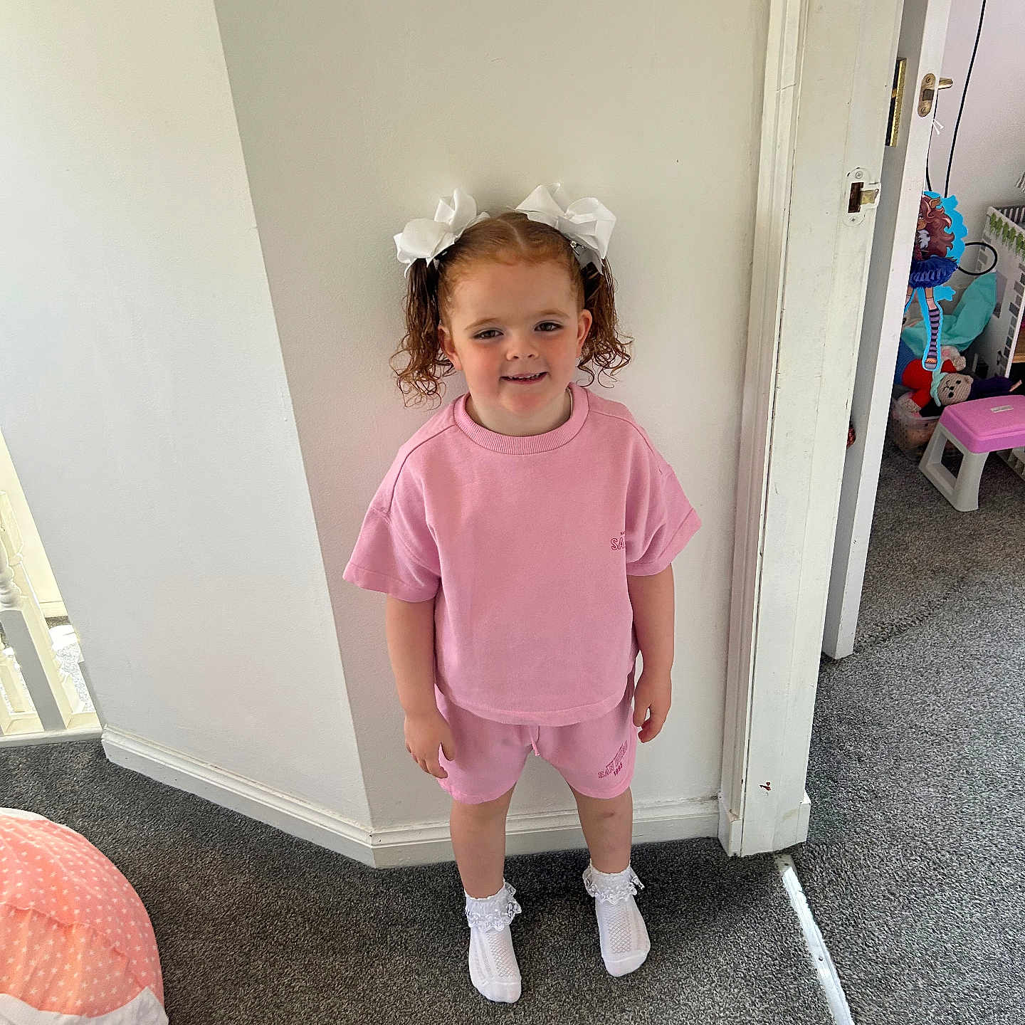 Freya May joined the competition — help win amazing prizes! carpet, casual_clothing, child, curly_hair, doorway, girl, happy, indoor, person, pink_outfit, playroom, shorts, smile, socks, standing, toy, tshirt, wall, white_bows, white_wall