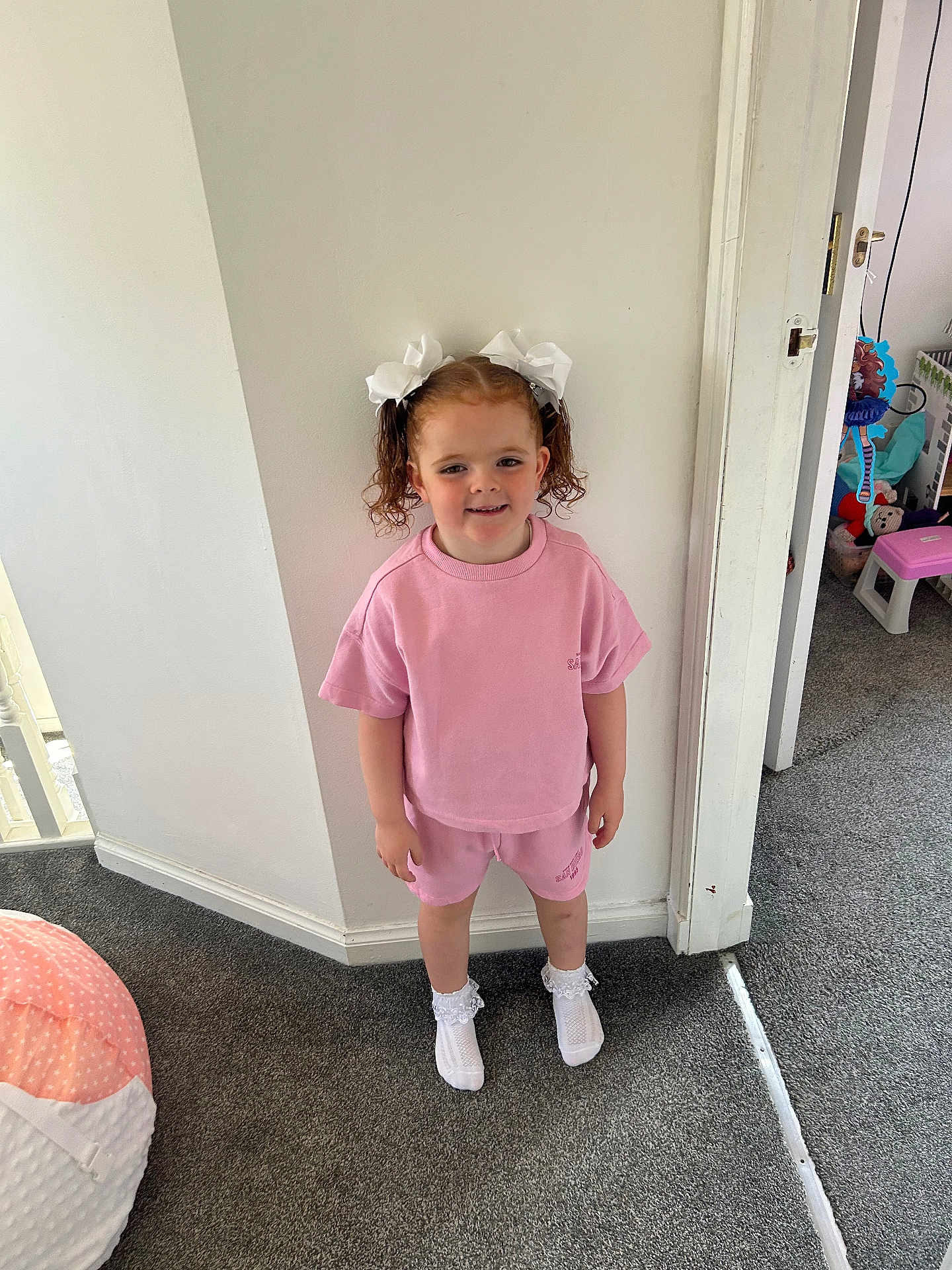 Freya May joined the competition — help win amazing prizes! child, girl, pink_outfit, white_bows, curly_hair, indoor, carpet, smile, socks, shorts, tshirt, standing, wall, doorway, toy, playroom, white_wall, casual_clothing, happy, person