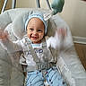 baby, child, smiling, blue_hat, baby_swing, cushion, indoor, happy, waving_arms, clothing, pants, seatbelt, sitting, face, person, cute, infant, playful, wood_floor, wall
