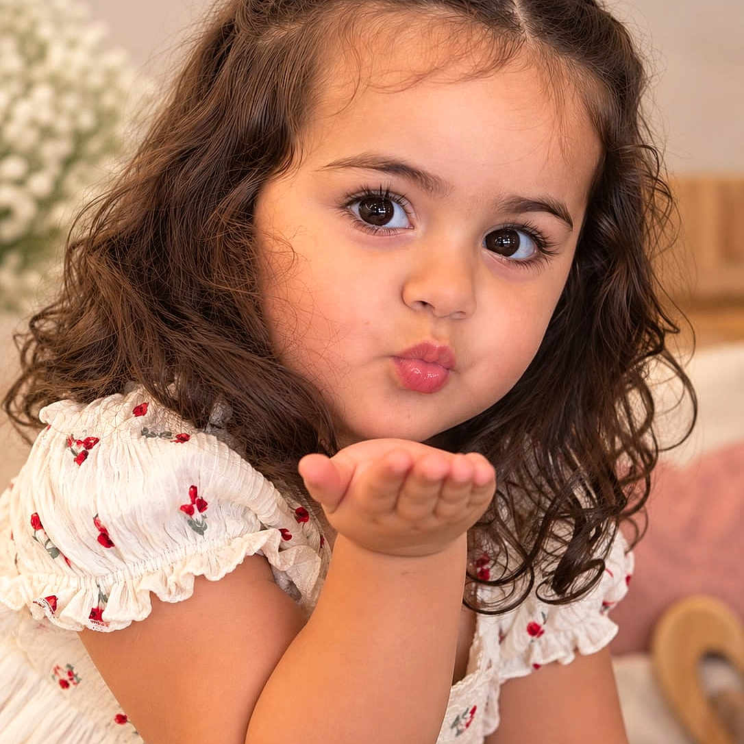 Gabriela is registered to the contest to win money with this photo: child, girl, portrait, curly_hair, brown_hair, hair_bow, smiling, blowing_kiss, hand, floral_dress, white_dress, indoors, seated, close_up, face, eyes, rosy_cheeks, wooden_chair, soft_lighting, photography