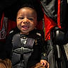 baby, infant, child, smiling, happy, face, portrait, car_seat, stroller, harness, safety_belt, seated, black_clothing, brown_pants, red_accent, outdoor, sunlight, close_up, vehicle, candid