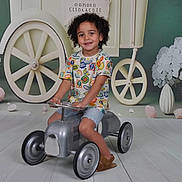 Charly participe au concours pour gagner de l'argent avec cette photo : child, toy_car, curly_hair, smiling, indoor, playful, colorful_clothing, shorts, shoes, floor, decor, flowers, wheels, background, happy, kid, portrait, cute, fun, young