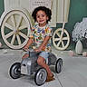 child, toy_car, curly_hair, smiling, indoor, playful, colorful_clothing, shorts, shoes, floor, decor, flowers, wheels, background, happy, kid, portrait, cute, fun, young