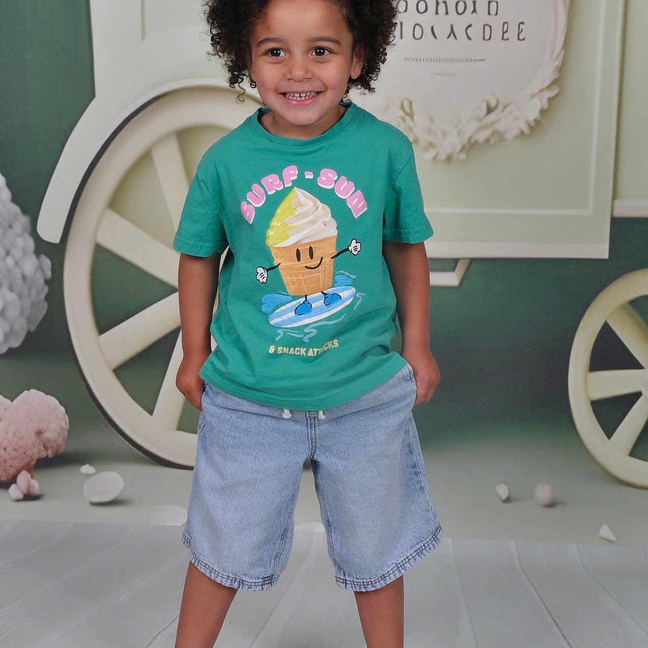 Charly participe au concours pour gagner de l'argent avec cette photo : child, boy, smiling, curly_hair, green_tshirt, denim_shorts, sandals, standing, indoor, background_props, cartwheel, floor, casual_clothing, happy, portrait, playful, cute, fashion, kids_fashion, studio