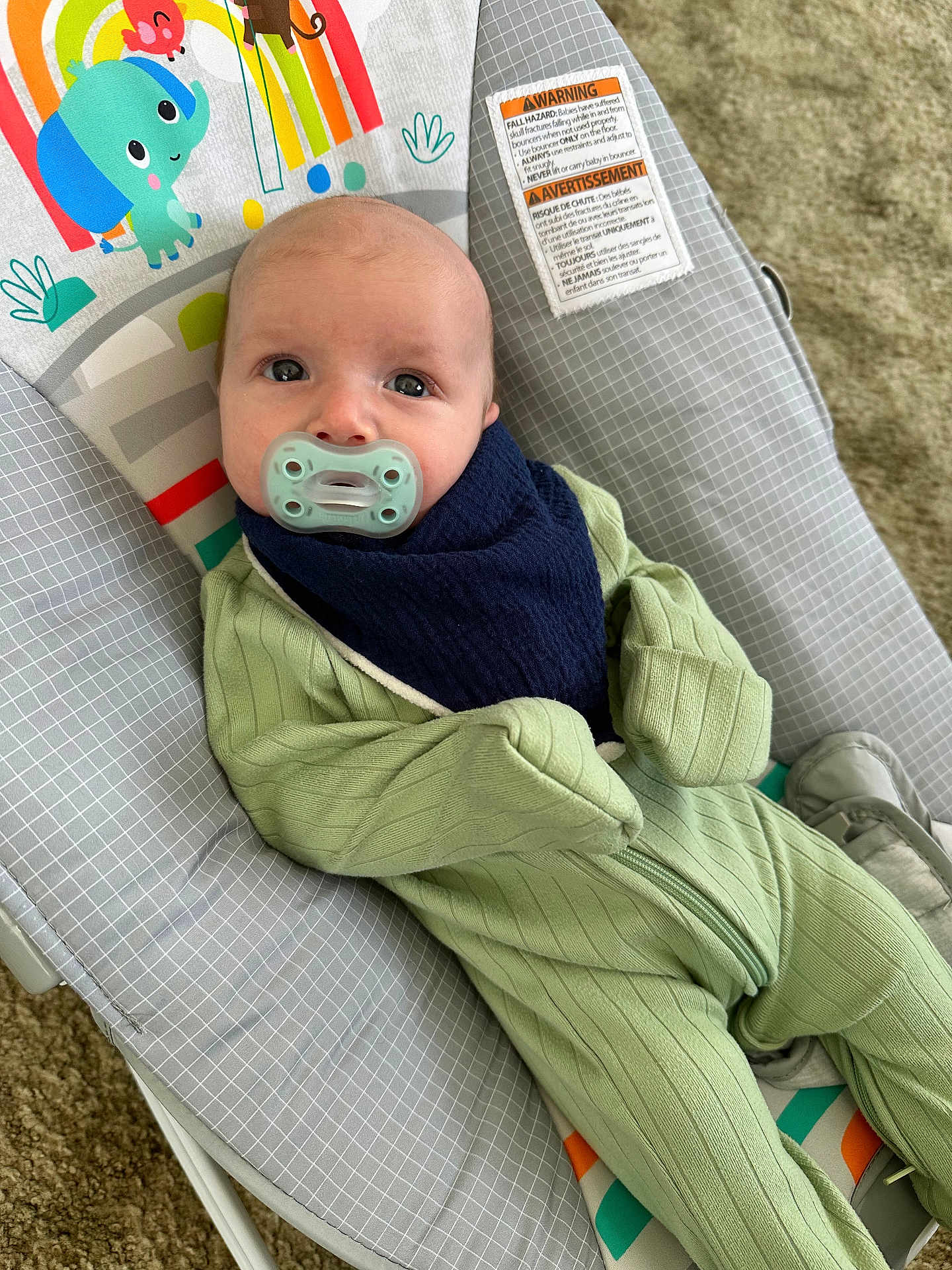 Teddy is registered to the contest to win money with this photo: baby, pacifier, onesie, bib, baby_seat, patterned, elephant, rainbow, colorful, carpet, infant, child, clothing, reclining, cute, indoors, soft, comfort, face, person