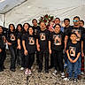 family, group, memorial, tents, flowers, black_tshirts, portrait, outdoor, adults, children, baby, men, women, casual_clothing, glasses, standing, gravel, somber, remembrance, diverse_ages