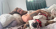 Porkchop is registered to the contest to win money with this photo: man, dog, bed, pillow, blanket, tattoo, smiling, cuddling, indoor, curtains, window, harness, resting, beard, hair, relaxed, person, animal, sleeping, home