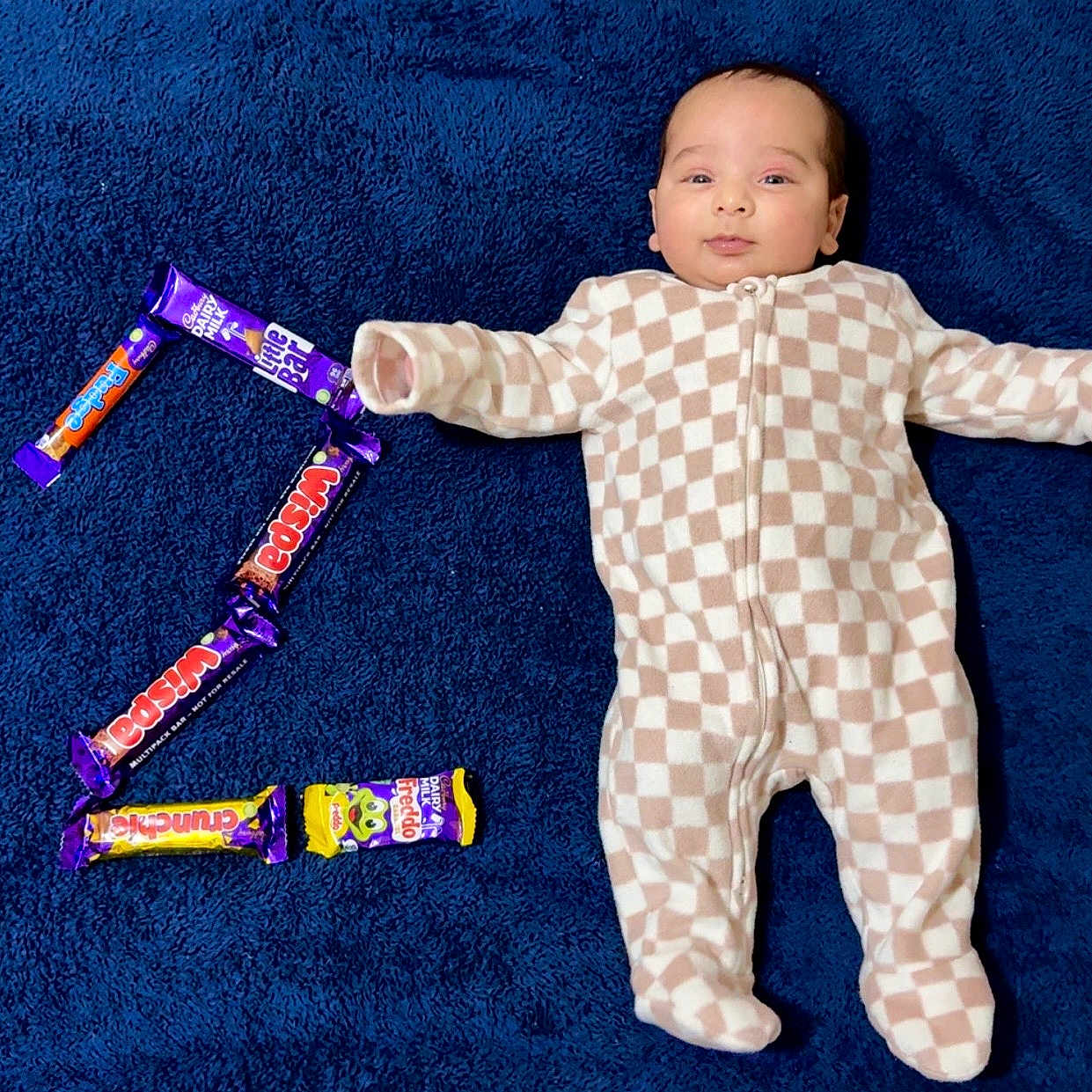 Manveer is registered to the contest to win money with this photo: baby, blue_blanket, candy_bars, checkered_pattern, child, comfortable, cute, face, indoor, infant, lying_down, number_two, onesie, portrait, relaxed, sleepwear, smiling, soft_texture, sweet_treats, young_child