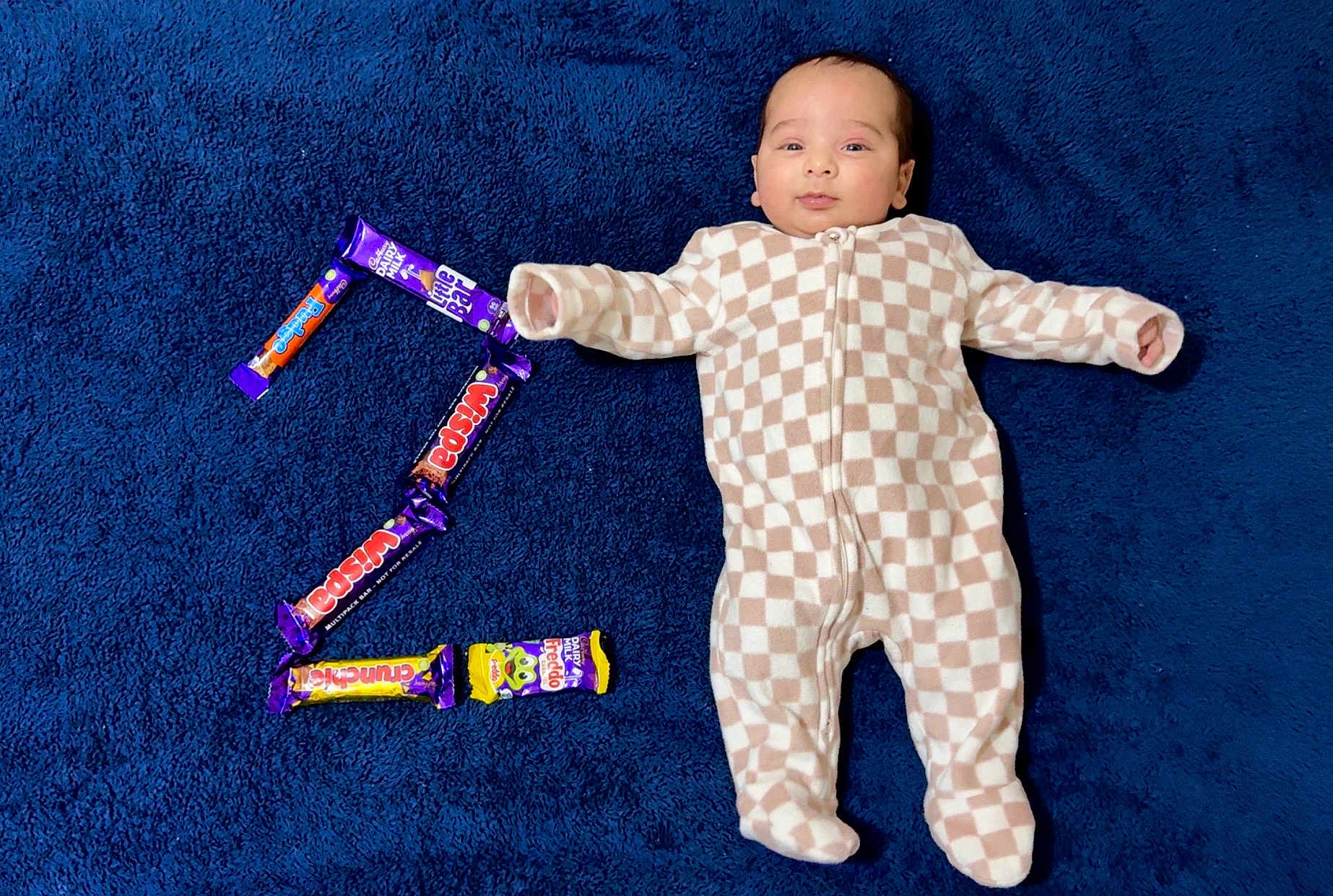 Manveer is registered to the contest to win money with this photo: baby, blue_blanket, candy_bars, checkered_pattern, child, comfortable, cute, face, indoor, infant, lying_down, number_two, onesie, portrait, relaxed, sleepwear, smiling, soft_texture, sweet_treats, young_child