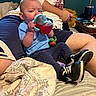 baby, child, sippy_cup, blue_shirt, toy, sneakers, bed, blanket, adult, shorts, hoodie, person, indoor, furniture, table, cup, candid, expression, clothing, casual