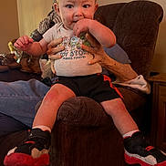 Micheal is registered to the contest to win money with this photo: toddler, child, baby, sitting, shoes, socks, toy_story_shirt, hands, armchair, furniture, indoor, person, face, legs, clothing, home, curious, supporting_hands, casual, cozy