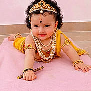 Veda joined the competition — help win amazing prizes! baby, child, cute, smiling, peacock_feather, jewelry, necklaces, bracelets, headgear, yellow_clothing, pink_blanket, floor, cultural, portrait, indoor, person, curly_hair, traditional, adorable, lying_down