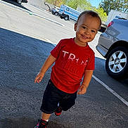 Julian is registered to the contest to win money with this photo: child, toddler, boy, smiling, outdoor, parking_lot, cars, vehicle, red_shirt, black_shorts, sneakers, walking, portrait, sunlight, daytime, asphalt, driveway, residential, playful, happy