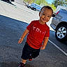 child, toddler, boy, smiling, outdoor, parking_lot, cars, vehicle, red_shirt, black_shorts, sneakers, walking, portrait, sunlight, daytime, asphalt, driveway, residential, playful, happy