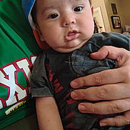 Mateo is registered to the contest to win money with this photo: baby, child, infant, cap, blue_cap, hand, person, adult_hand, clothing, green_shirt, indoor, living_room, furniture, curious, cute, portrait, people, holding, casual, home