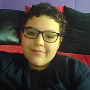 Logan is registered to the contest to win money with this photo: child, glasses, curly_hair, smile, sofa, cushion, indoor, portrait, casual, relaxed, young_person, black_shirt, red_cushion, black_cushion, purple_wall, face, person, seated, home, comfortable