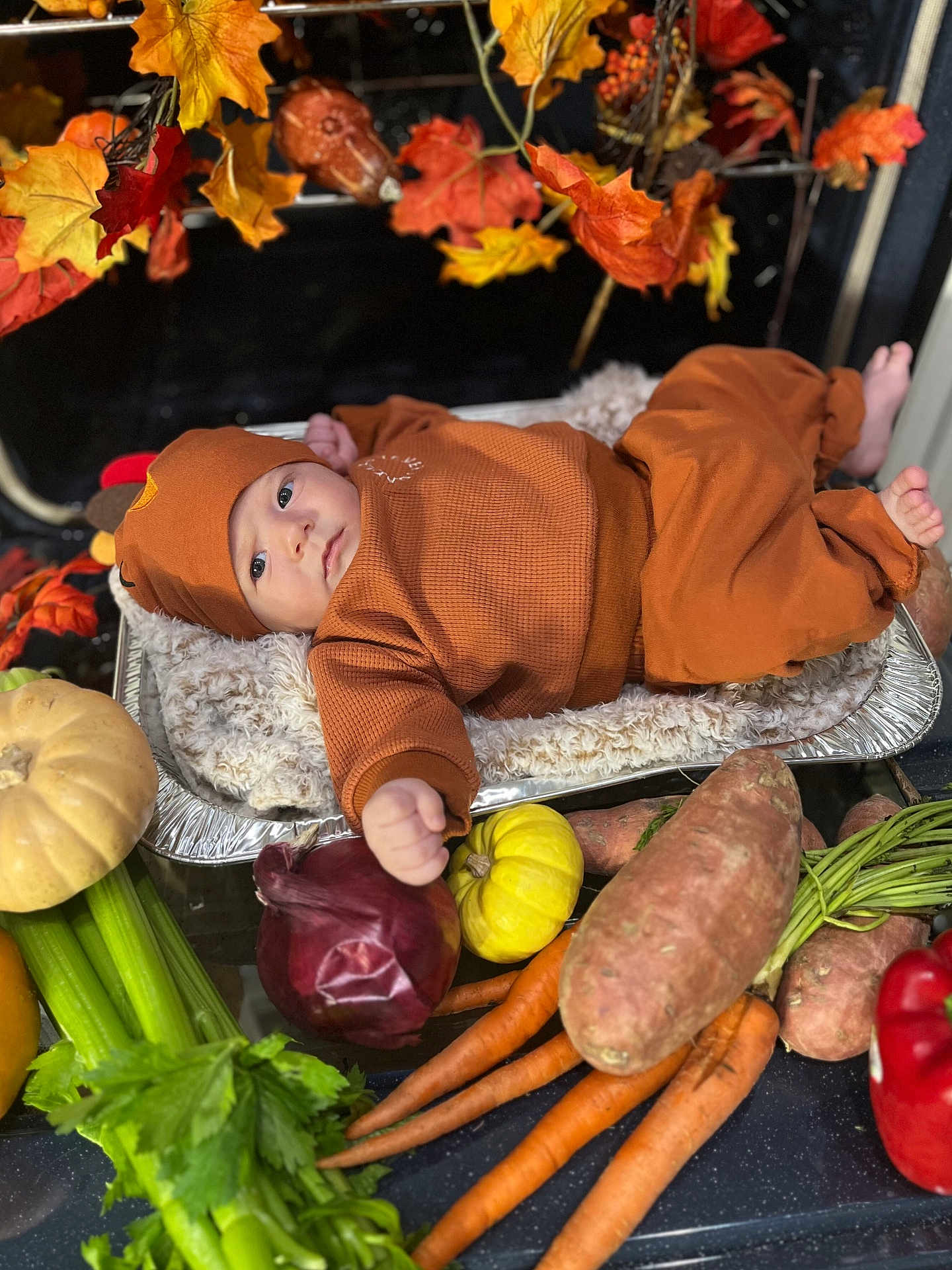 Gideon is registered to the contest to win money with this photo: baby, infant, brown_clothing, blanket, foil_tray, vegetables, carrots, celery, sweet_potatoes, red_onion, yellow_pumpkin, red_bell_pepper, fall_leaves, autumn, cozy, soft_texture, indoor, curious, lying_down, still_life