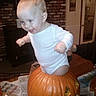 baby, child, pumpkin, halloween, carved_pumpkin, indoor, onesie, smiling, happy, face, person, hands, floor, newspaper, brick_wall, furniture, door, decor, head, playful