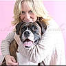 Dakota is registered to the contest to win money with this photo: woman, dog, boxer_dog, hugging, smiling, blonde_hair, pink_background, portrait, indoor, happy, pet, animal, friendship, affection, warm_clothing, earrings, face, closeup, cuddling, love