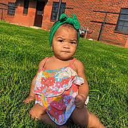 Nyomi is registered to the contest to win money with this photo: baby, child, toddler, portrait, outdoor, grass, lawn, green_bow, headband, floral_outfit, dress, sitting, building, brick_wall, sunlight, face, expression, infant, fashion, photography