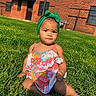 baby, child, toddler, portrait, outdoor, grass, lawn, green_bow, headband, floral_outfit, dress, sitting, building, brick_wall, sunlight, face, expression, infant, fashion, photography