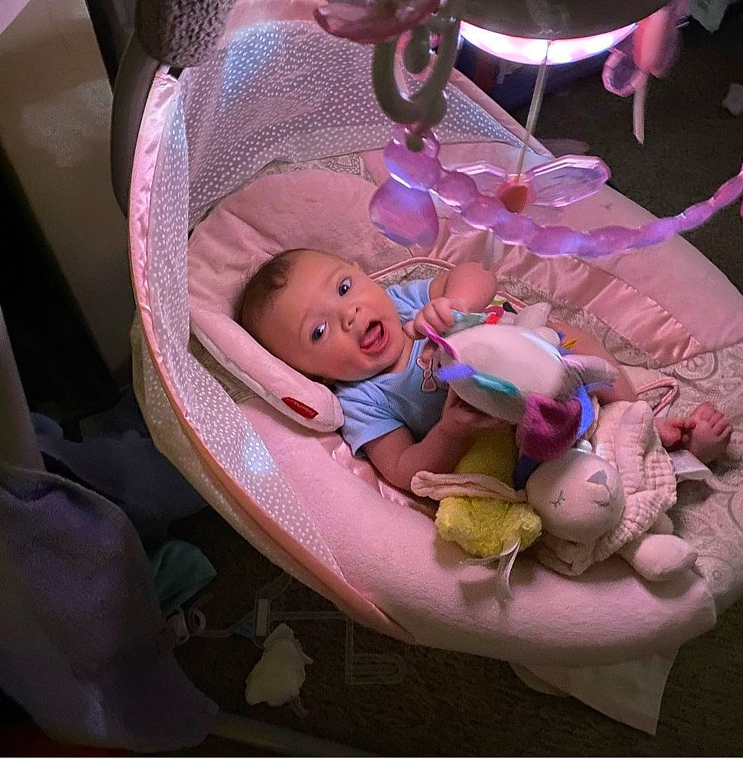 Everest is registered to the contest to win money with this photo: baby, baby_products, baby_sleeping, baby_toddler_clothing, cheek, child, comfort, fashion_accessory, fun, happy, headgear, lap, linens, magenta, person, pink, purple, sitting, smile, surprise