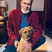 Chance joined the competition — help win amazing prizes! man, dog, puppy, indoor, floor, beard, red_shirt, kneeling, wooden_wall, lamp, couch, chain_necklace, window_blinds, hand, pet, brown_dog, casual_clothing, portrait, home, warm_lighting