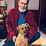 man, dog, puppy, indoor, floor, beard, red_shirt, kneeling, wooden_wall, lamp, couch, chain_necklace, window_blinds, hand, pet, brown_dog, casual_clothing, portrait, home, warm_lighting