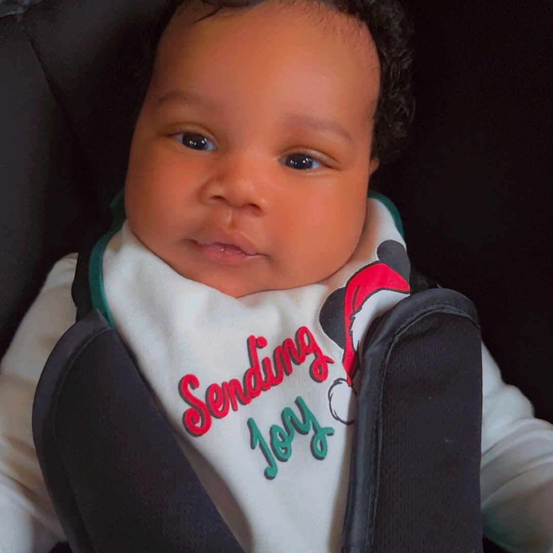 Jacques is registered to the contest to win money with this photo: baby, bib, black_hair, car_seat, child, closeup, clothing, curly_hair, cute, face, holiday_theme, indoors, infant, joy, person, portrait, seat_belt, sitting, smile, young_child