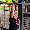 child, boy, playground, playground_equipment, blue_rings, swing, smiling, outdoor, trees, grass, wood_chips, black_shirt, shorts, wristband, fun, active, daylight, park, happy, leisure
