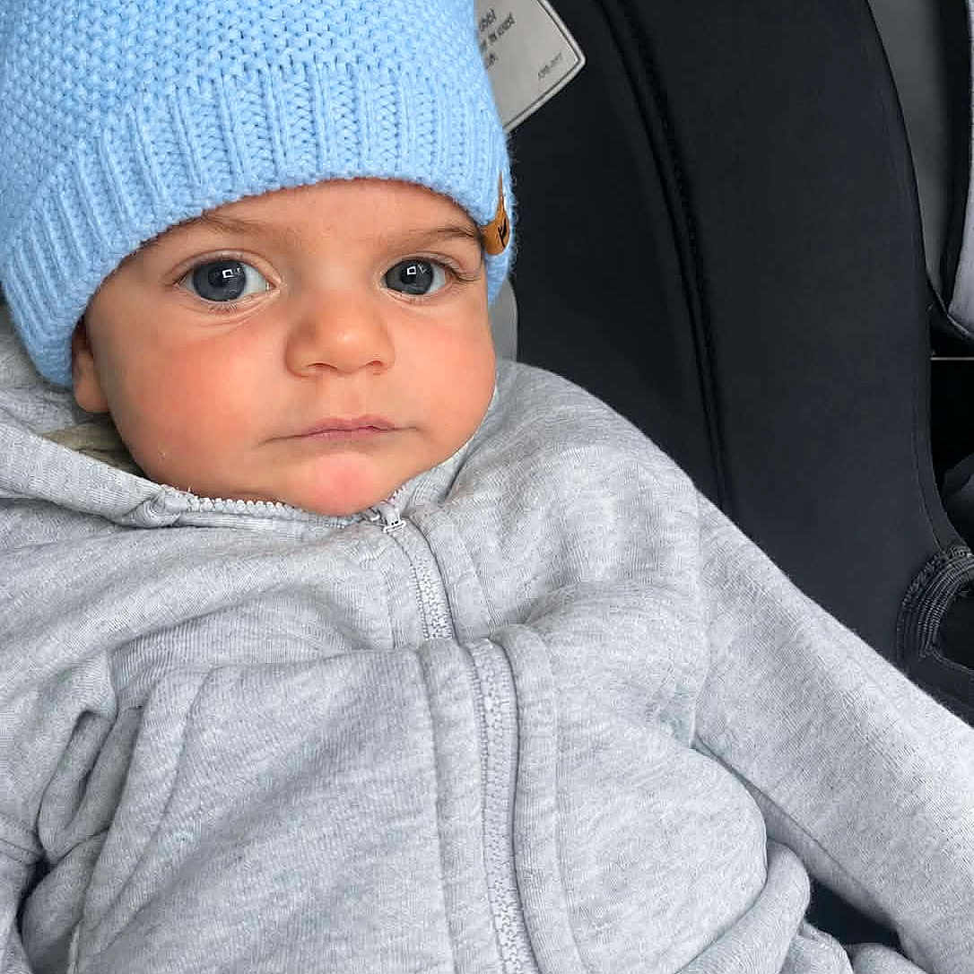 Adam Mostafa is registered to the contest to win money with this photo: baby, child, infant, car_seat, car, blue_hat, knit_hat, gray_clothing, jacket, zipper, portrait, face, eyes, serious_expression, sitting, indoors, close_up, travel, cozy, photography