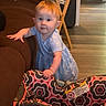 baby, child, toddler, standing, blue_dress, curious, face, portrait, indoors, living_room, couch, car_seat, furniture, folding_table, wood_floor, home, baby_gear, colorful_pattern, smiling, photography