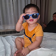 Ethan joined the competition — help win amazing prizes! toddler, child, sunglasses, orange_shirt, bed, blanket, indoors, person, adult, phone, curtains, bracelet, smiling, cute, playful, casual, home, seated, foot, hair