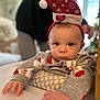 baby, child, infant, christmas_hat, holiday, festive, santa_hat, sweater, cute, smiling, face, hands, indoor, cozy, cushion, holiday_season, person, portrait, reaching, soft_focus