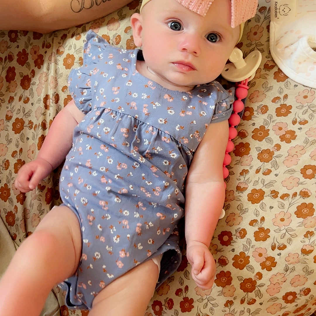 Saileigh joined the competition — help win amazing prizes! arm, baby, bedspread, blue_onesie, bow, child, closeup, cute, expression, floral_pattern, headband, indoors, infant, lying_down, pacifier, person, skin, soft_lighting, tattoo, toy