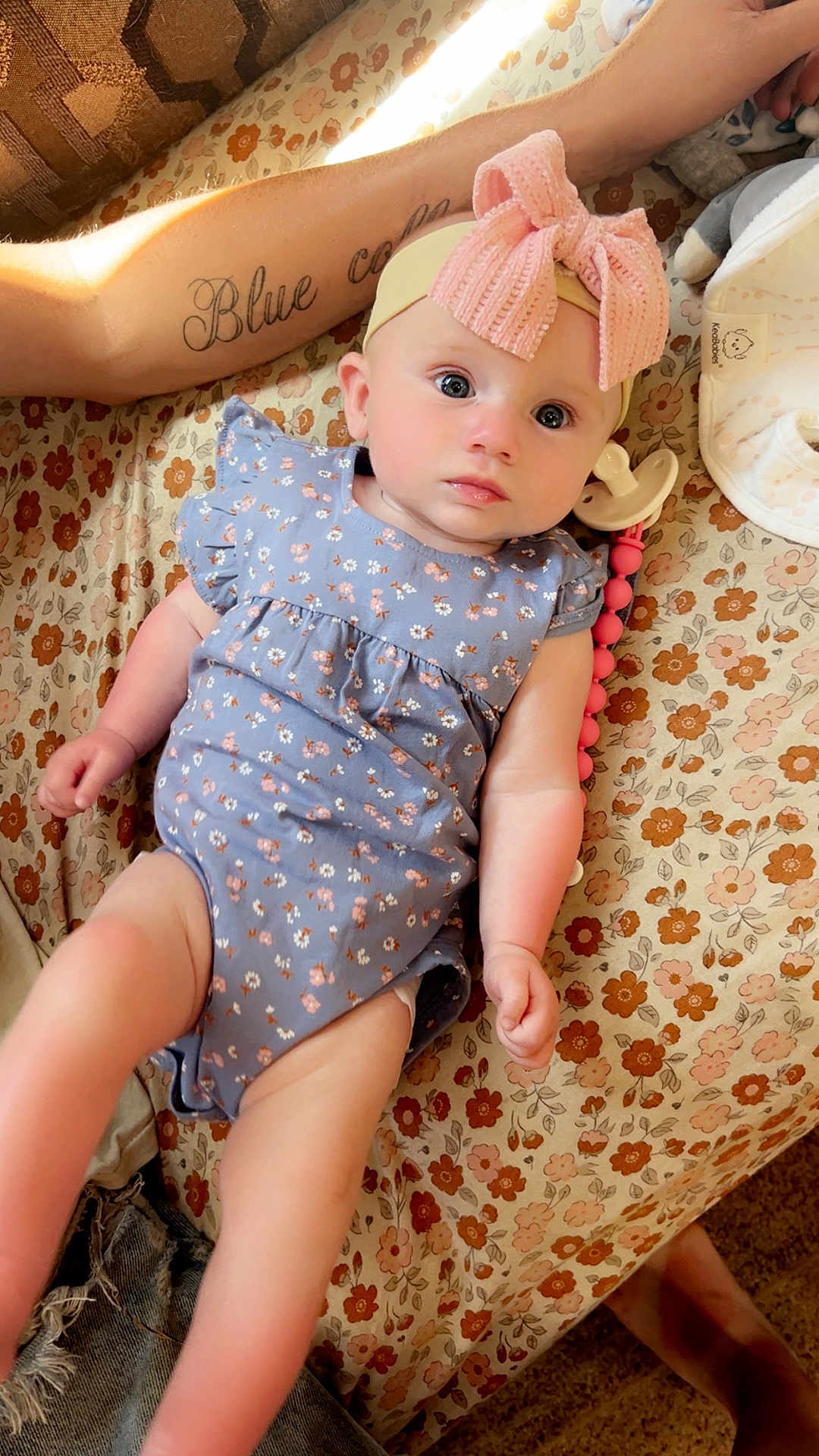 Saileigh joined the competition — help win amazing prizes! baby, infant, headband, bow, blue_onesie, floral_pattern, tattoo, arm, bedspread, pacifier, toy, skin, person, cute, lying_down, indoors, soft_lighting, expression, closeup, child