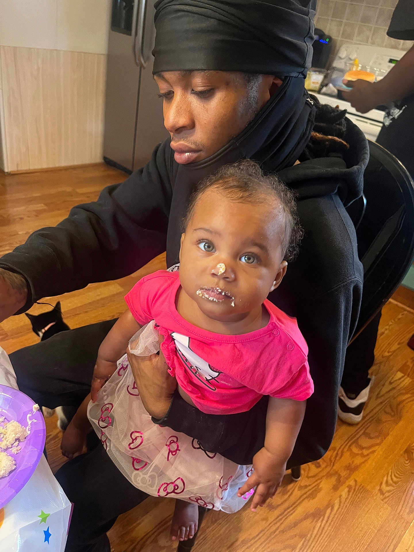 Aaliyah is registered to the contest to win money with this photo: baby, child, person, adult, cake, frosting, kitchen, food, table, plate, floor, cat, clothing, tutu, pink_shirt, sitting, holding, indoor, wooden_floor, curly_hair