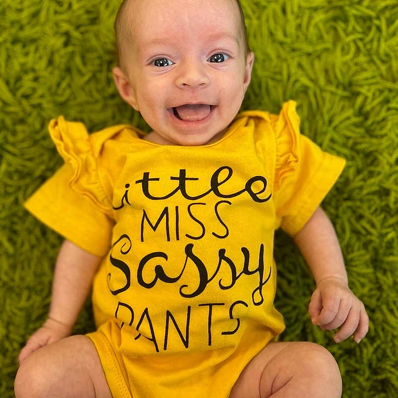 Eden is registered to the contest to win money with this photo: baby, child, smiling, yellow_onesie, text_on_clothing, green_rug, happy, infant, cute, portrait, laying_down, playful, human, face, skin, expression, sitting, candid, indoors, one_person