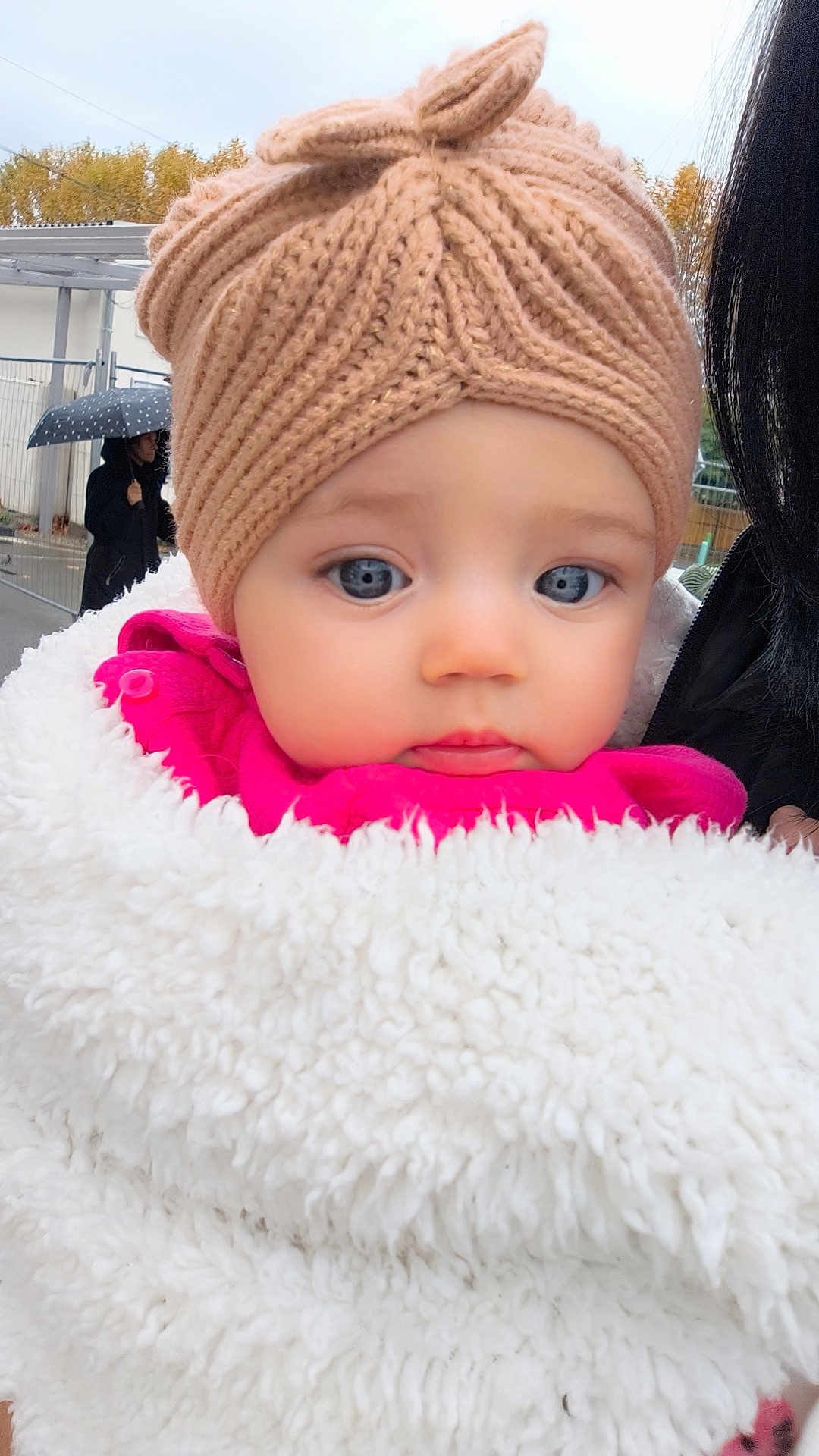 Esmée participe au concours pour gagner de l'argent avec cette photo : baby, child, portrait, face, blue_eyes, knitted_hat, beige_hat, blanket, white_blanket, pink_clothing, outdoor, rainy_day, umbrella, person, winter_clothing, cute, close_up, warm, soft_texture, curious