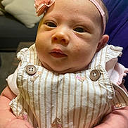 Avalon is registered to the contest to win money with this photo: baby, infant, headband, flower, overalls, striped_clothing, hands, person, indoor, couch, pillow, closeup, cute, skin, face, portrait, child, newborn, soft_lighting, holding