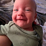 James is registered to the contest to win money with this photo: baby, smiling, face, onesie, green_clothing, indoor, blanket, happy, infant, portrait, cute, child, newborn, person, cozy, soft_light, closeup, head, skin, expression