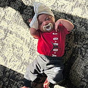 Naomi is registered to the contest to win money with this photo: baby, infant, pacifier, headband, bow, red_shirt, jordan, nike_pants, carpet, patterned_rug, sunlight, shadow, indoor, floor, clothing, cute, relaxed, small_feet, laying_down, child