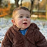 child, toddler, outdoor, autumn, snow, brown_jacket, fuzzy_clothing, cute, curious, face, portrait, nature, trees, blurred_background, winter_clothing, young_child, expression, sitting, seasonal, warm_clothes