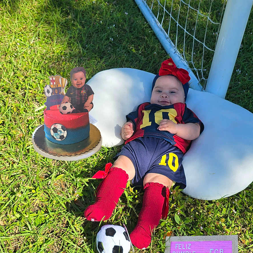 Aisha Sophie is registered to the contest to win money with this photo: baby, birthday, blue_shorts, cake, celebration, child, goal_net, grass, letter_board, number_10, outdoor, photography, pillow, red_bow, red_socks, smile, soccer, sportswear, sunlight, toy_ball