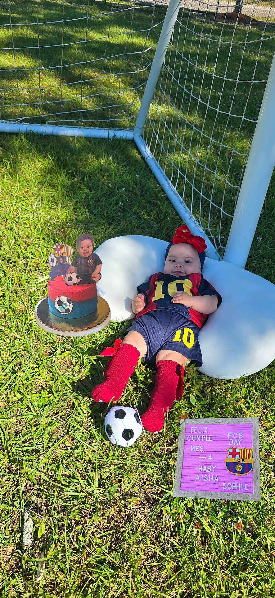 Aisha Sophie is registered to the contest to win money with this photo: baby, soccer, grass, outdoor, goal_net, cake, pillow, red_bow, red_socks, number_10, celebration, birthday, letter_board, toy_ball, blue_shorts, sunlight, child, smile, sportswear, photography