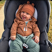 Isaac is registered to the contest to win money with this photo: baby, child, stroller, hoodie, bear_ears, green_pants, smiling, happy, outdoor, sidewalk, grass, cute, infant, clothing, casual_wear, portrait, seated, baby_face, playful, warm_clothes