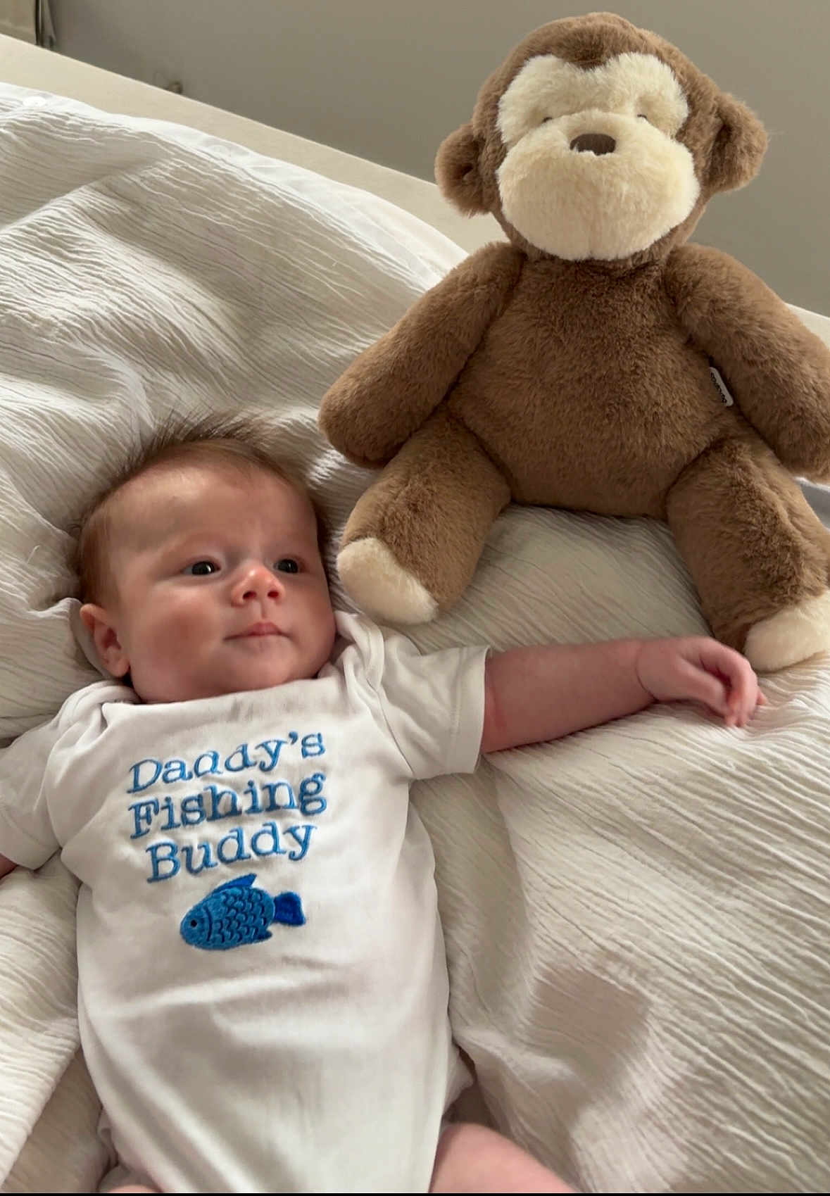 Reuben is registered to the contest to win money with this photo: baby, infant, onesie, plush_toy, monkey_toy, bed, blanket, cute, child, soft_toy, smiling, indoor, resting, white_clothing, adorable, cozy, sleepwear, fabric, lying_down, young_child