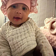 Dream is registered to the contest to win money with this photo: baby, infant, pink_bonnet, white_bib, cozy, blanket, indoor, person, portrait, cute, child, soft_lighting, window_blinds, sleepwear, closeup, smiling, young_child, headwear, warm_clothing, bedroom
