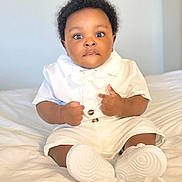 Rhian is registered to the contest to win money with this photo: baby, child, infant, white_clothing, sitting, bed, curly_hair, wide_eyes, fists, cute, portrait, indoor, soft_lighting, young_child, footwear, person, adorable, expression, skin, face